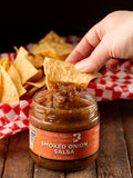Butterfly Bakery of Vermont Smoked Onion Salsa jar with a tortilla chip being dipped into it, on a wooden surface.