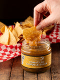 Tortilla chip being dipped into a jar of Tomatillo Chipotle salsa from Butterfly Bakery of Vermont on a wood surface.
