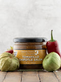 Jar of Tomatillo Chipotle Salsa from Butterfly Bakery of Vermont surrounded with red jalapenos and tomatillos on a wooden surface