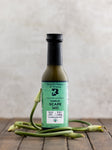 A bottle of Garlic Scape Sauce, displayed on a wooden surface with several garlic scapes to the side.