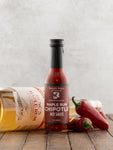 Bottle of Maple Rum Chipotle hot sauce on a wooden surface with peppers and a bottle of rum in the background.