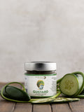 A jar of Gustard mustard with a label featuring the band Guster's logo, surrounded by fresh cucumbers and jalapeno peppers.